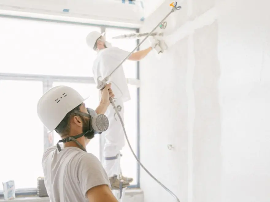 Plasterers in masks and hard hats spraying plaster on an interior wall
