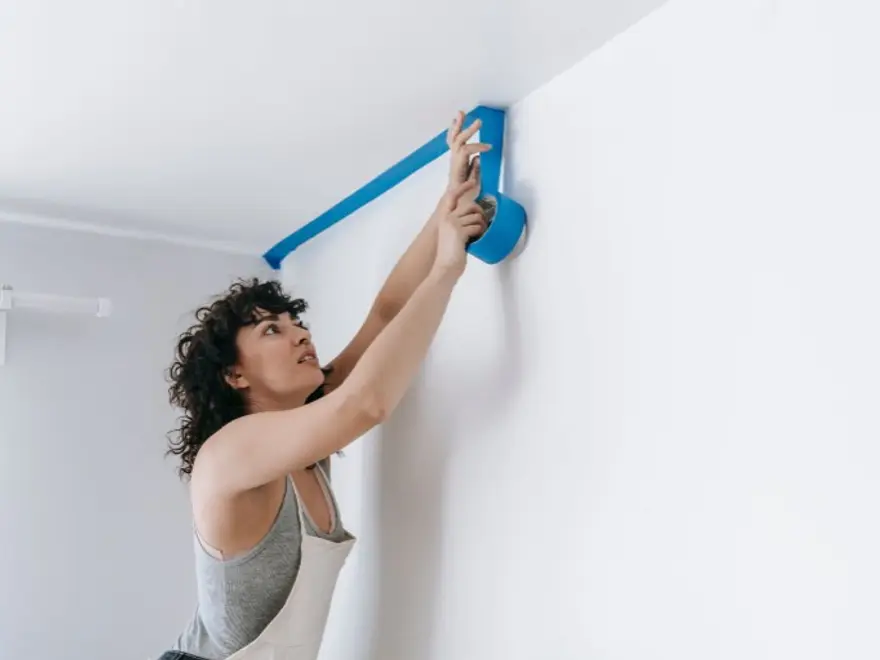 Decorator applying blue tape along the edge of a ceiling